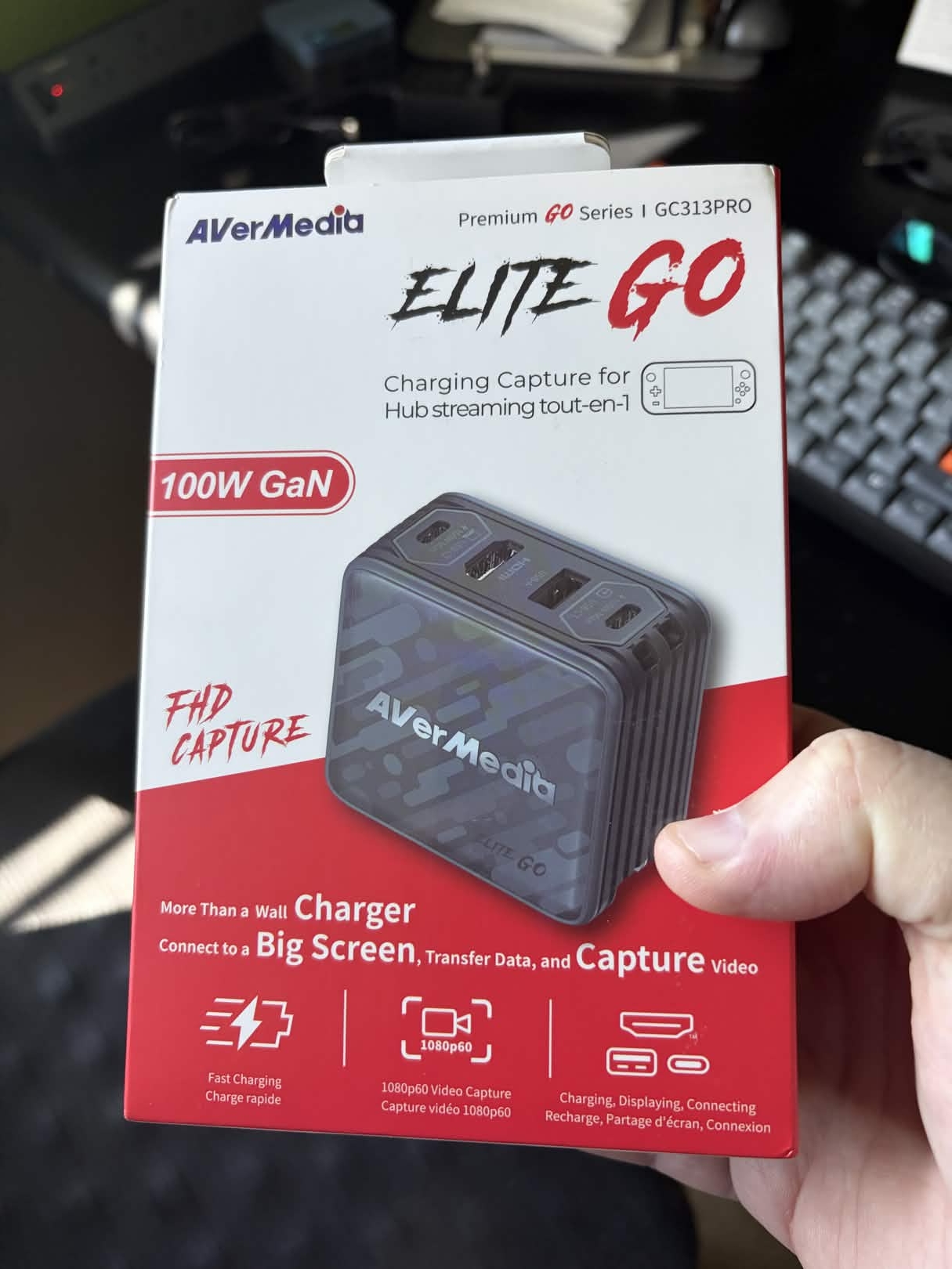 Box for an AVerMedia Elite Go charging capture device, a hub that connects to a big screen, transfers data, and captures video, with the device displayed on the box. The box reads: 'AVerMedia, Premium GO Series | GC313PRO, ELITE GO, Charging Capture for, Hub streaming tout-en-1, 100W GaN, FHD CAPTURE, More Than a Wall Charger, Connect to a Big Screen, Transfer Data, and Capture video, Fast Charging, 1080p60 Video Capture, Charging, Displaying, Connecting'.