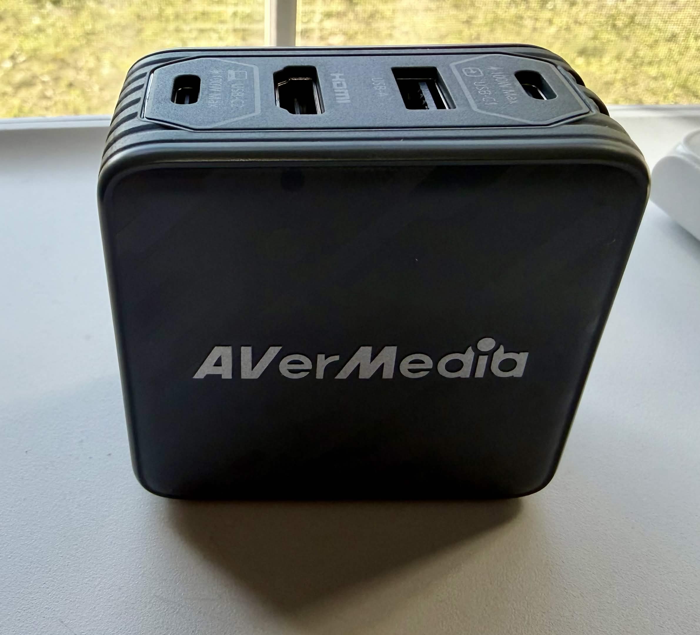 Black AVerMedia cube-shaped device with ports and the brand name printed on the front. On the top are the words: "USB-C", "HDMI", and "USB-C", "100W Max" printed on the ports.